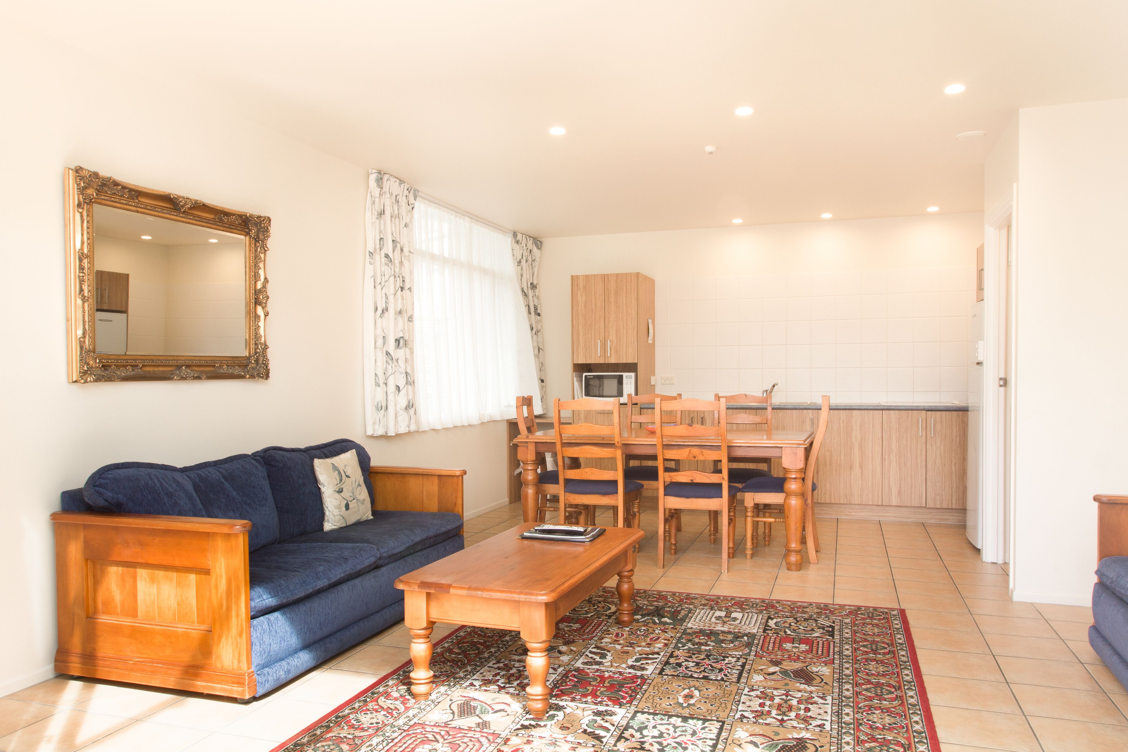 A living room with a sofa, a wooden coffee table, a patterned rug, a dining area with a table and chairs, and a kitchen in the background.