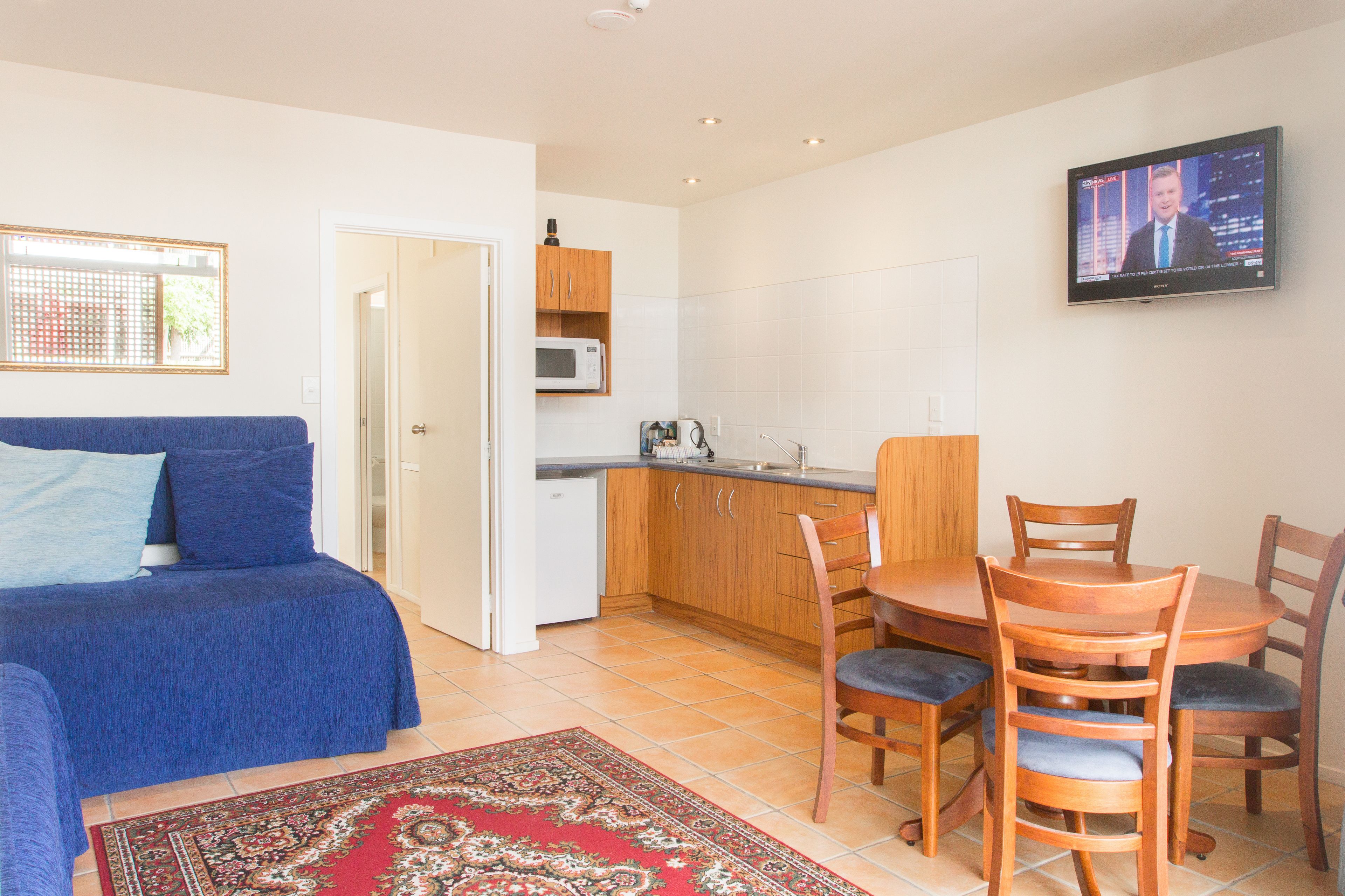 A compact living space with a blue sofa, a dining table with chairs, a kitchen area, and a TV mounted on the wall.