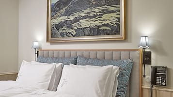 Una cama bien tendida con sábanas blancas y almohadas estampadas, un cuadro de paisaje enmarcado encima, una lámpara de noche y un teléfono sobre una mesita de noche de madera.