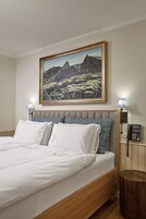 Una cama bien tendida con sábanas blancas y almohadas estampadas, un cuadro de paisaje enmarcado encima, una lámpara de noche y un teléfono sobre una mesita de noche de madera.