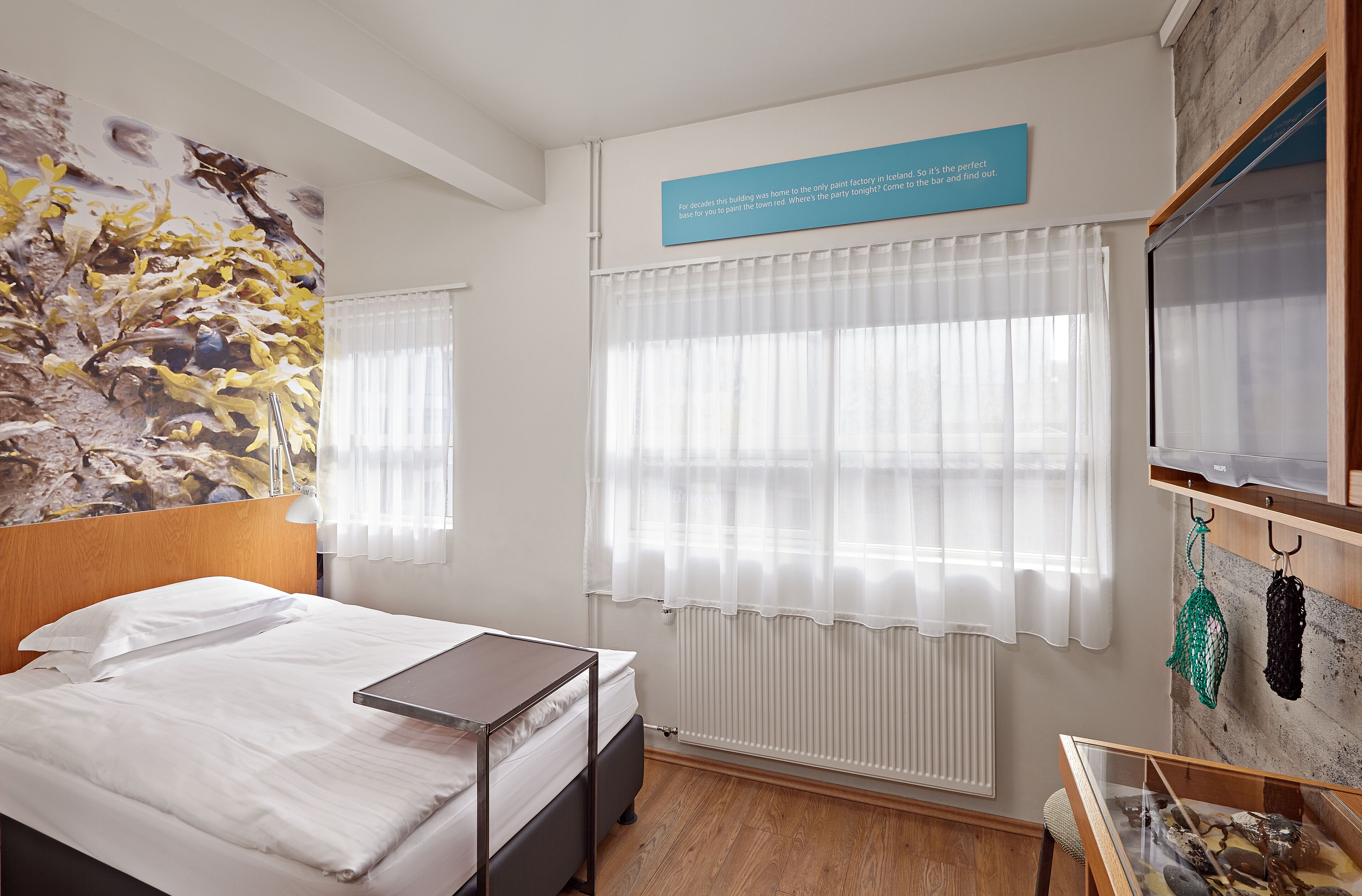 A hotel room with a bed, a desk, a radiator, and a wall with a painting.