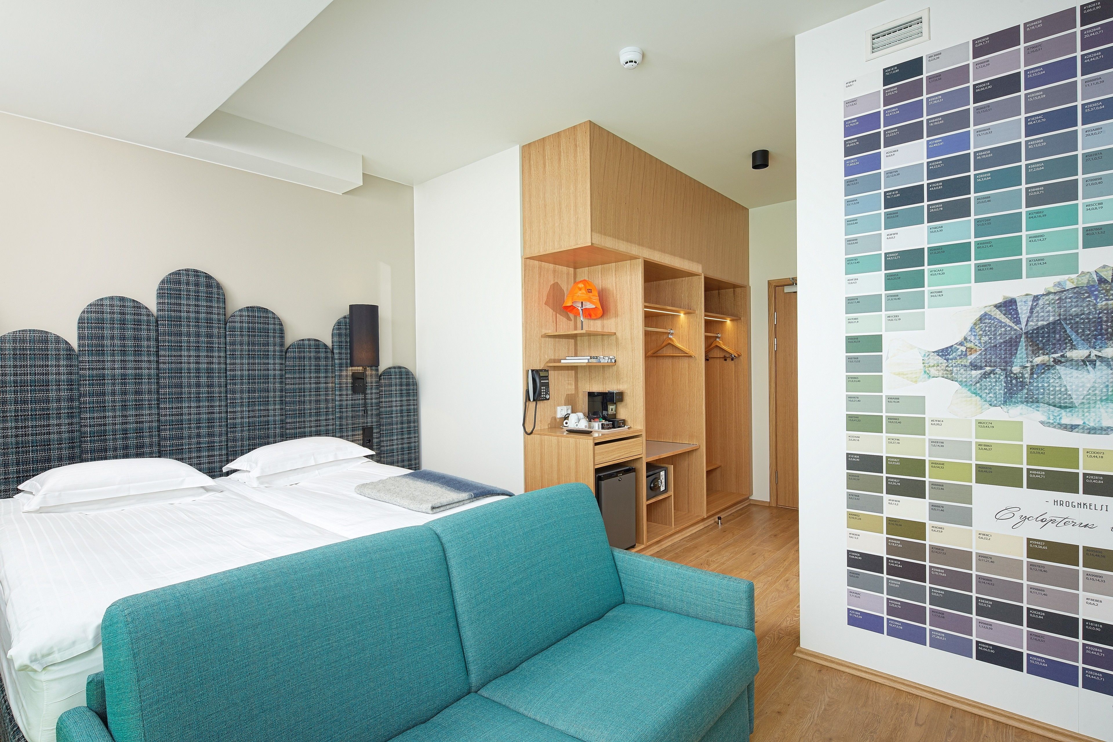 Ein Hotelzimmer mit Bett, einem petrolfarbenen Sofa, einem Holzschrank, einer kleinen Küchenzeile und einer Farbmusterkarte an der Wand.