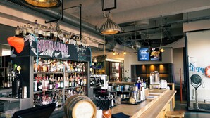 Uma área de bar com prateleiras de garrafas de bebidas, um caixa registradora e banquetas.