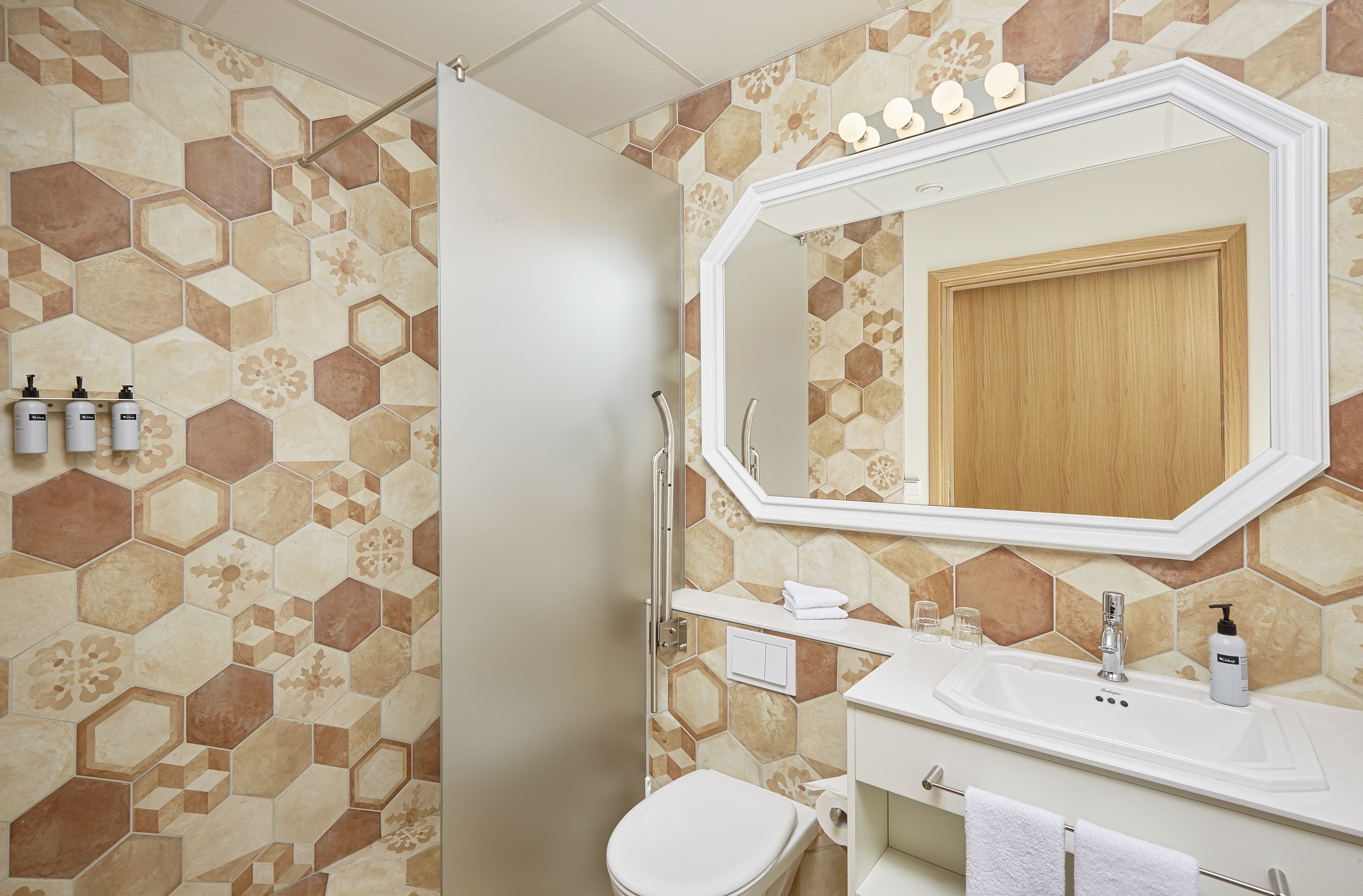 Een badkamer met een betegelde muur in een honingraatpatroon, een grote spiegel, een wit wastafelmeubel en een toilet.