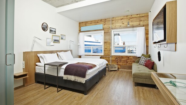 Quarto moderno com uma cama grande, um sofá e vista para um navio pelas janelas.