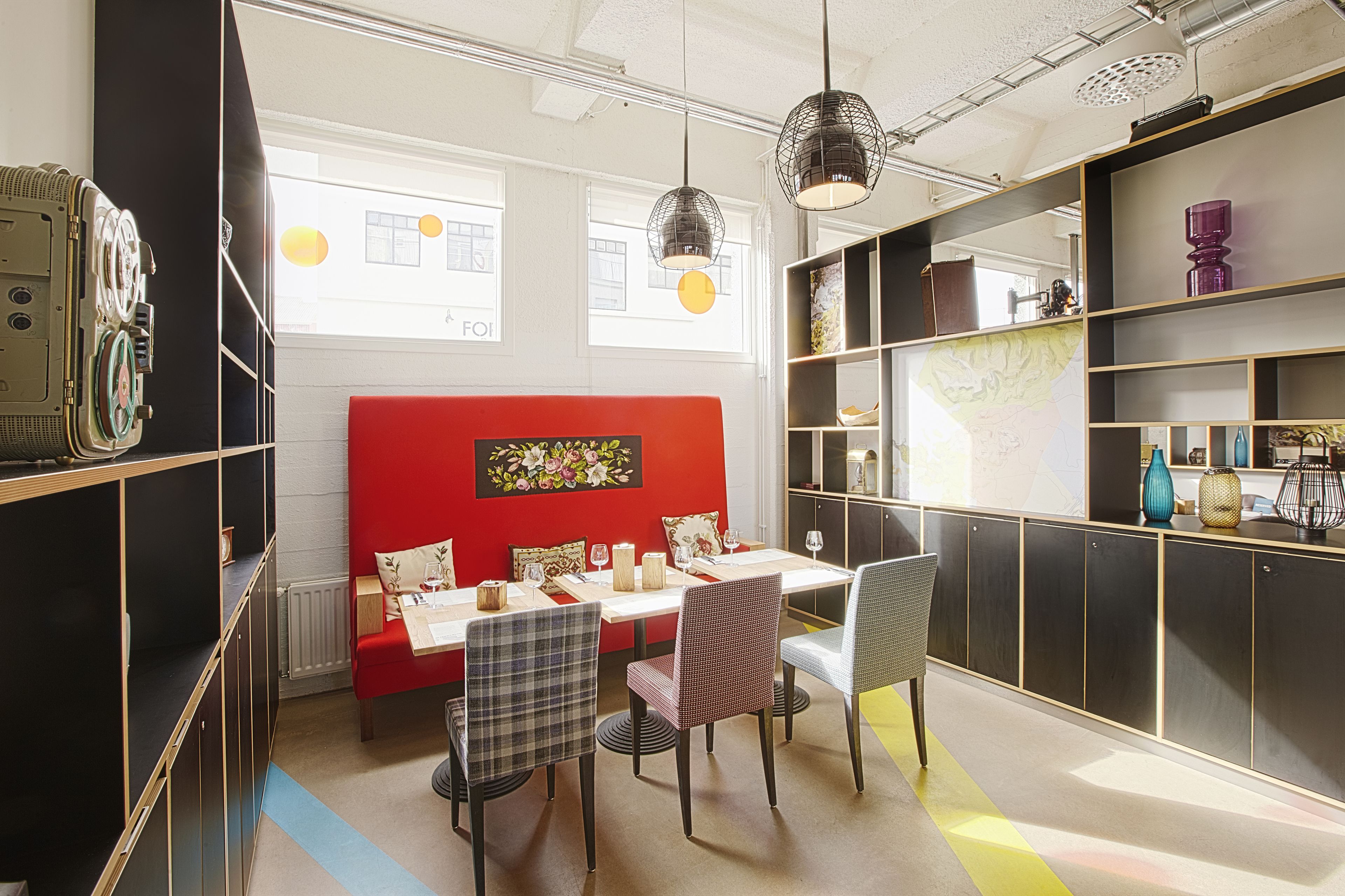 A dining area with a red booth, a white table, and black chairs. There are shelves with decorative items and a large window with natural light.