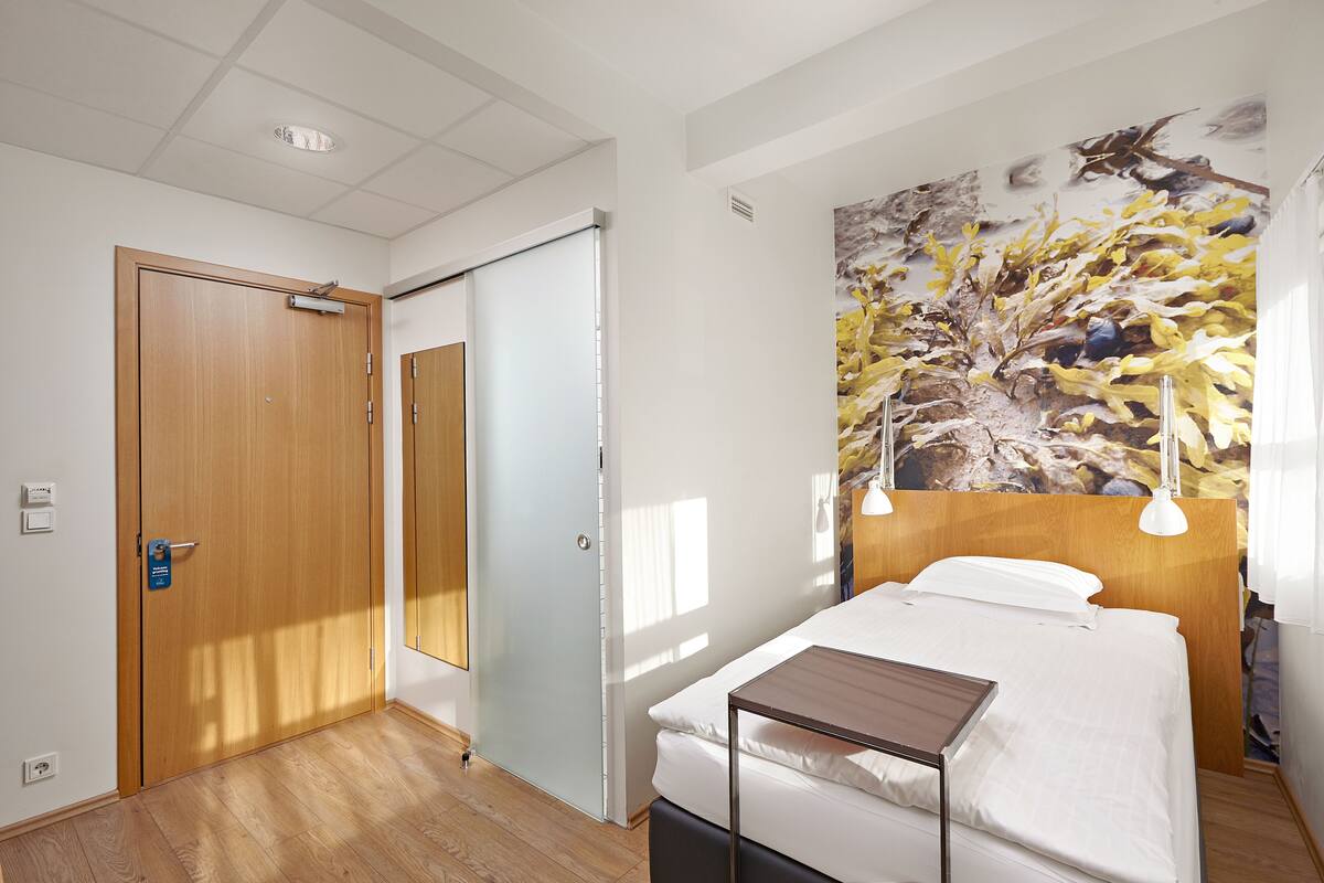 Een hotelkamer met een bed, een klein tafeltje, een houten deur en een groot abstract schilderij aan de muur.