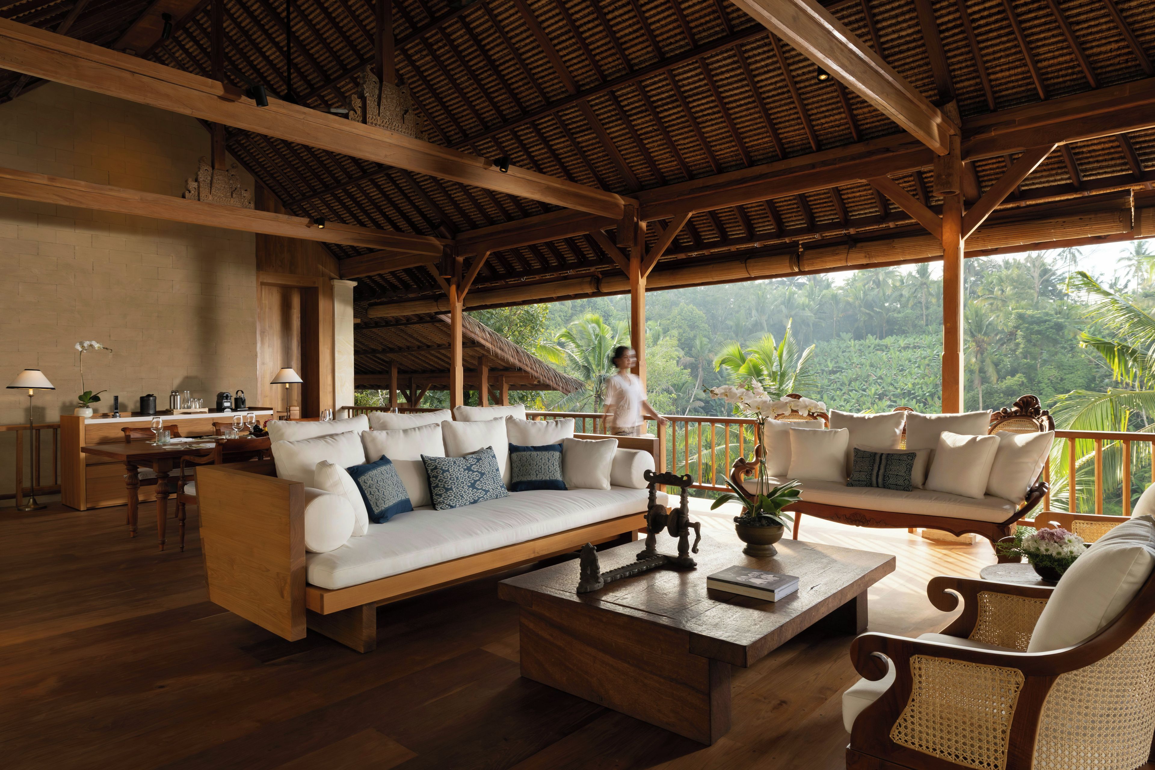 A spacious living area with a wooden ceiling, a sofa set, a dining table, and a view of lush greenery outside.