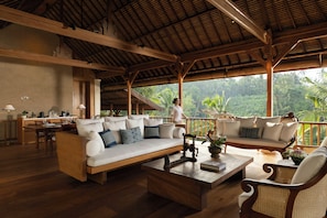 A spacious living area with a wooden ceiling, a sofa set, a dining table, and a view of lush greenery outside. - COMO Shambhala Estate (Ubud)