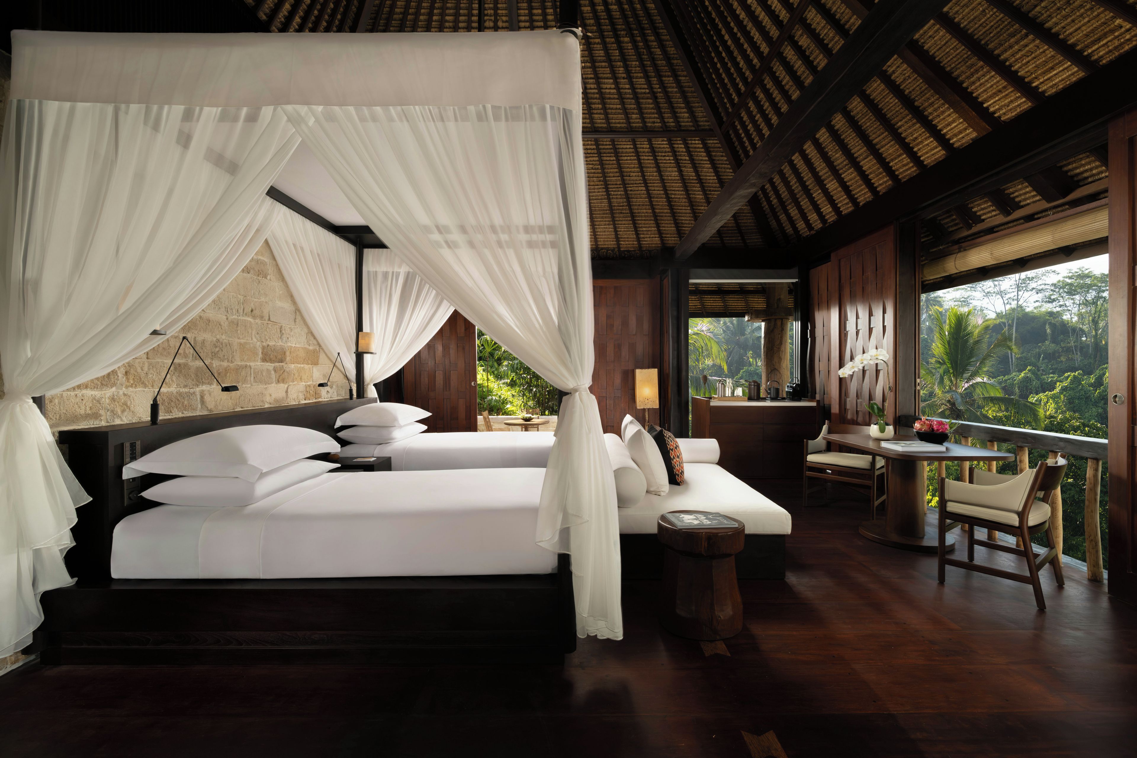 A four-poster bed with white linens and a canopy, a wooden table with chairs, and a view of the surrounding greenery.
