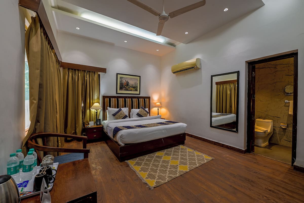 Habitación de hotel con cama, mesitas de noche, escritorio, silla, baño con inodoro y espejo, y unidad de aire acondicionado.