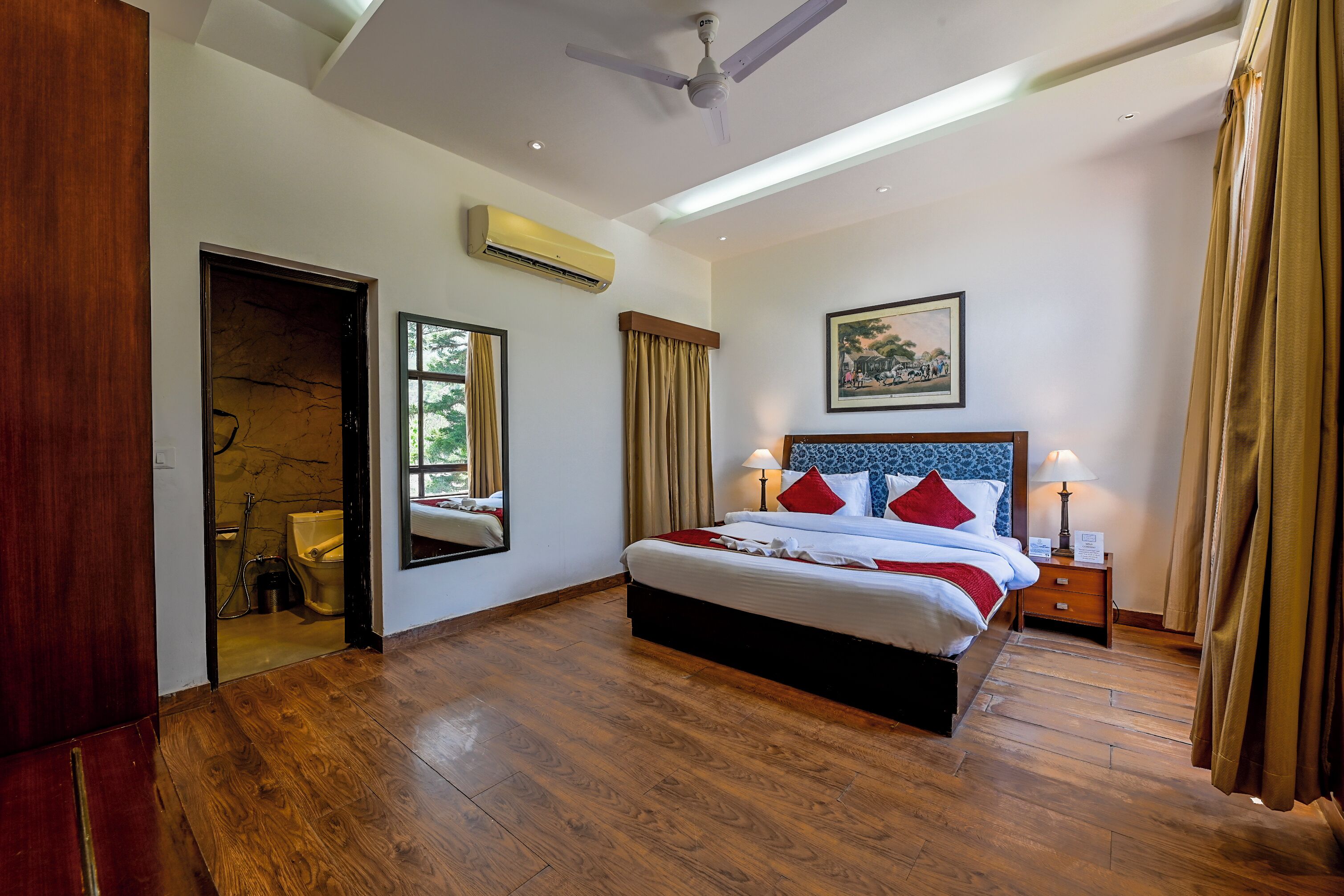 Habitación de hotel con piso de madera, cama con ropa de cama roja y blanca, mesita de noche con lámpara, ventilador de techo y unidad de aire acondicionado.