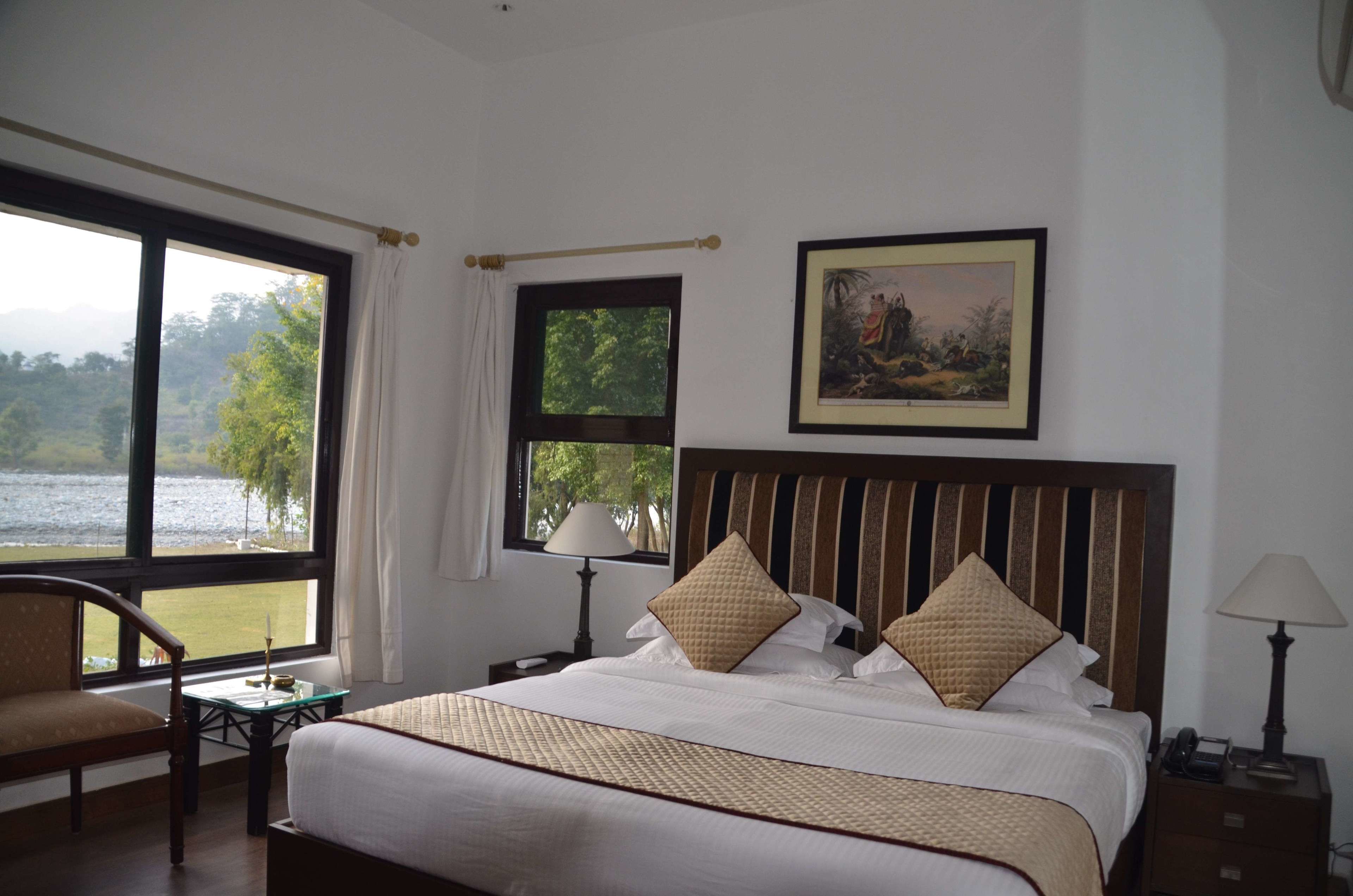 A hotel room with a large bed, a chair, a table, a nightstand, a lamp, a window with a view of a lake, and a framed picture on the wall.