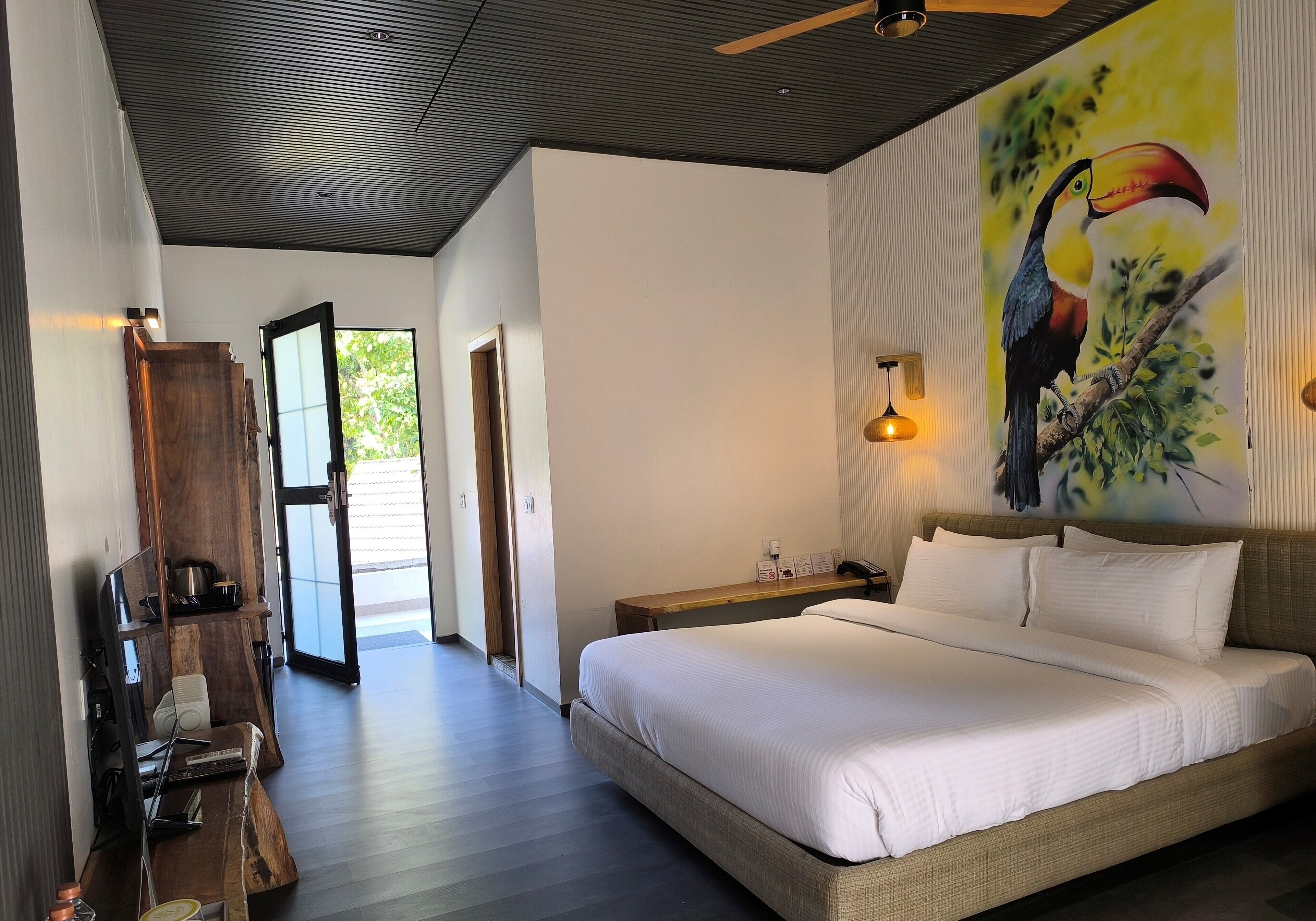 Une chambre avec un grand lit, un bureau en bois, un ordinateur portable et un tableau représentant un toucan accroché au mur.