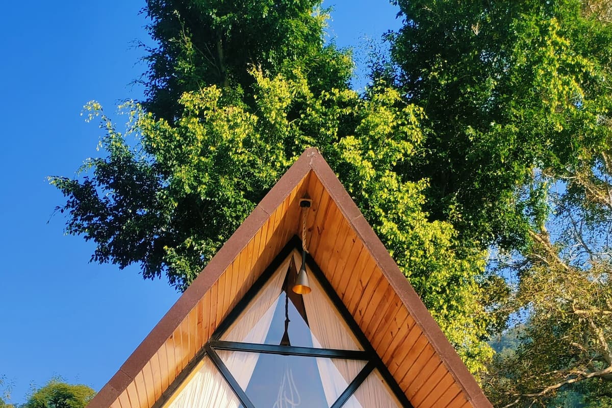 Una cabaña de madera con techo triangular, ventanas amplias y una campana colgando del ápice.