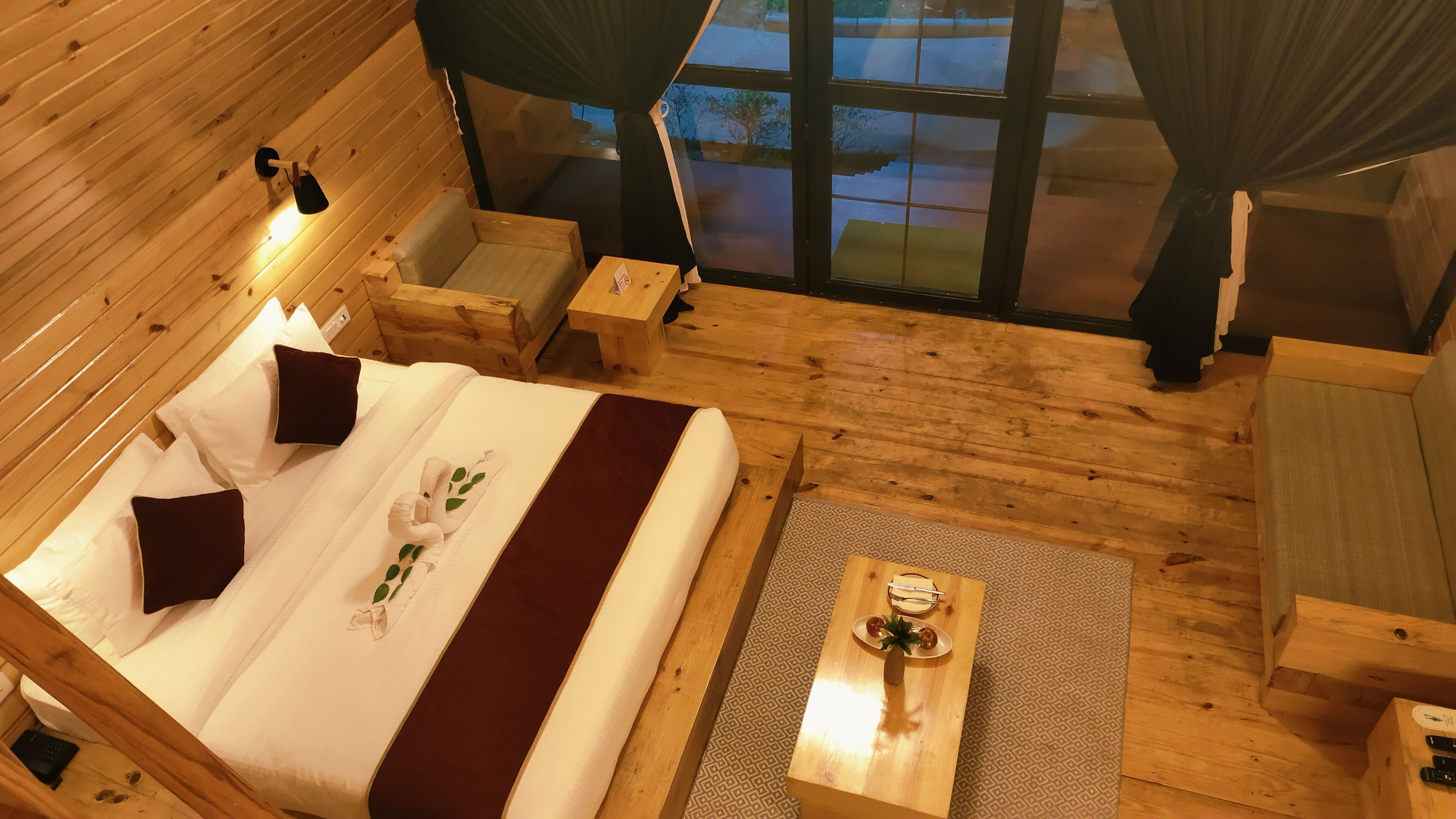 Una habitación tipo cabaña de madera con una cama, una mesita con una piña, y vista al exterior por una ventana.