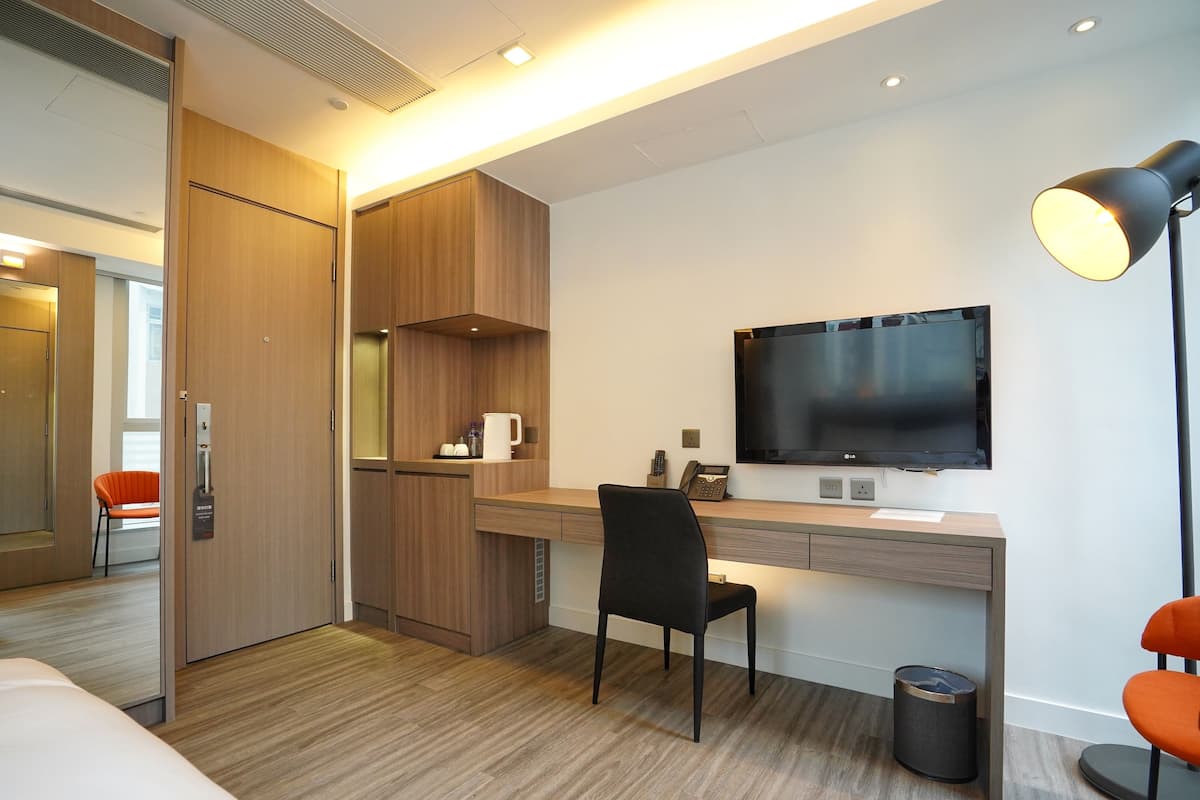 A hotel room with a wooden desk, a black chair, a television, and a lamp.