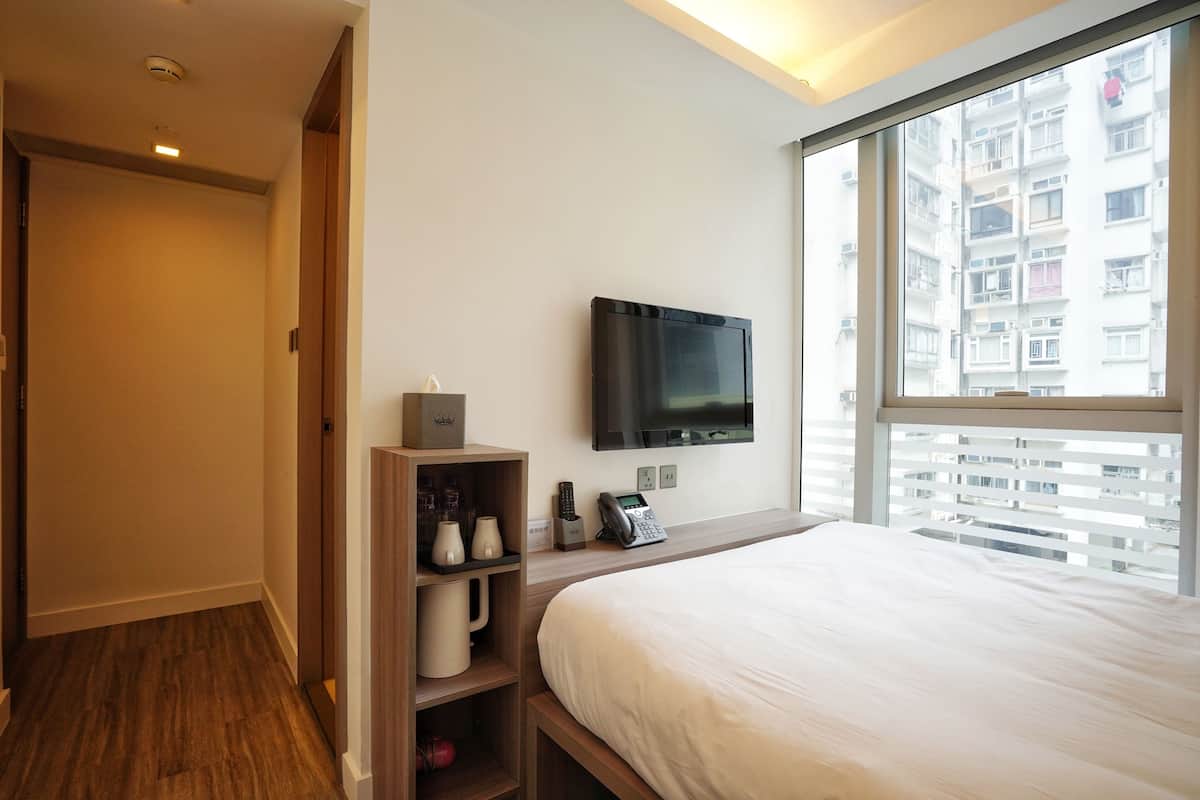 A hotel room with a large bed, a flat-screen TV mounted on the wall, a wooden shelf with toiletries, and a window with a city view.