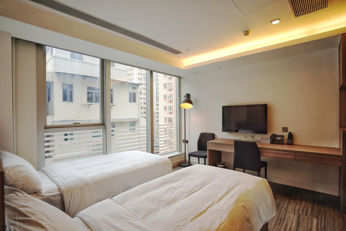 A hotel room with two beds, a desk, a chair, a TV, and a large window with a view of buildings.