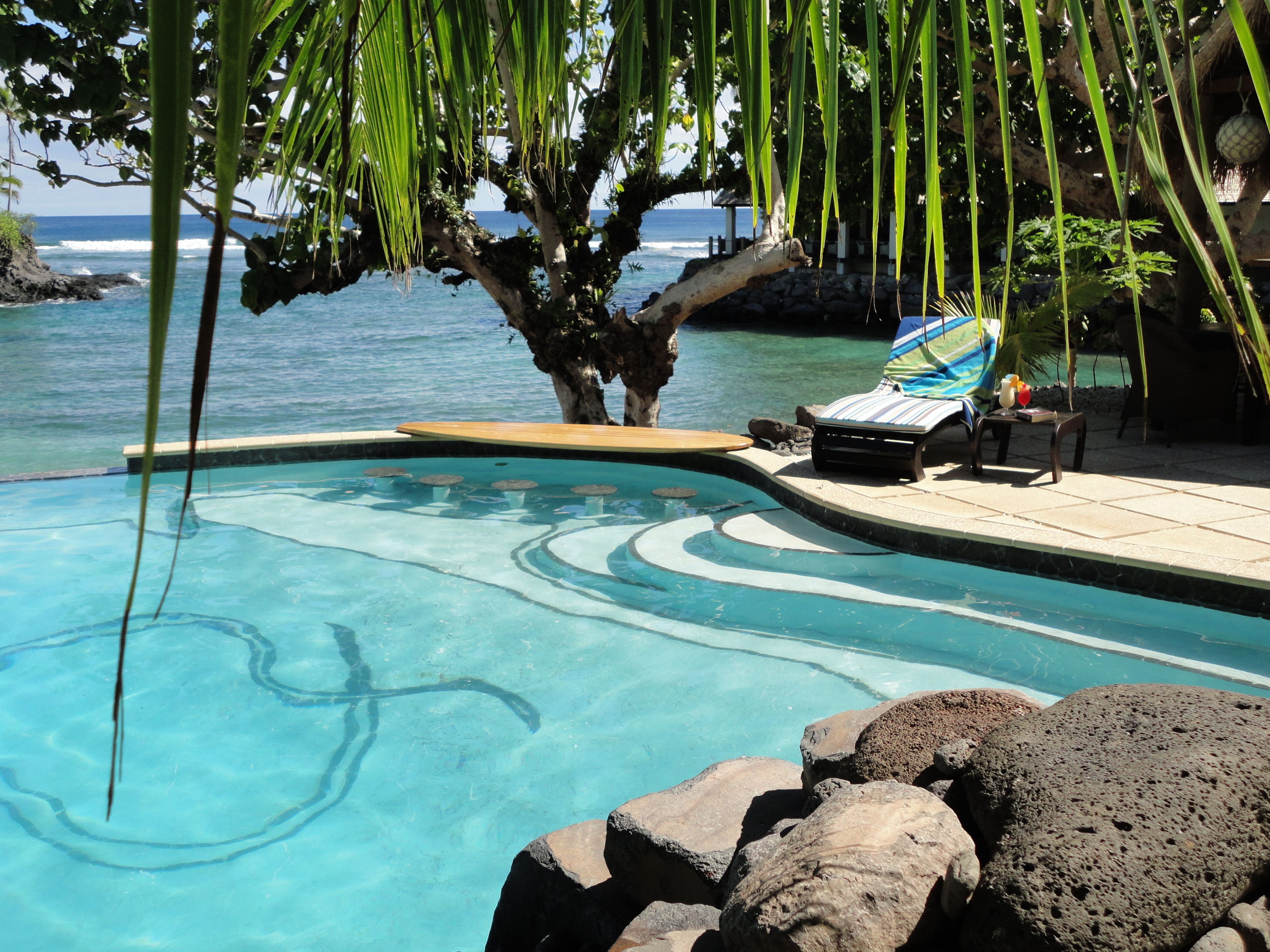 Ein klarer Pool mit geschwungener Kante, umgeben von Steinen und einer Liege mit gestreiftem Handtuch.