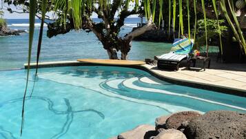 Ein klarer Pool mit geschwungener Kante, umgeben von Steinen und einer Liege mit gestreiftem Handtuch.