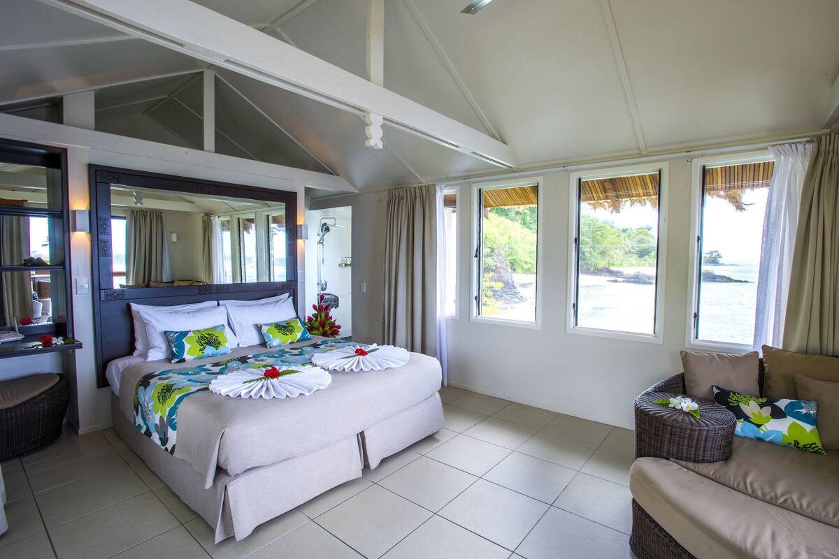 A hotel room with a large bed, a sofa, a small table, and a window with a view of the sea.