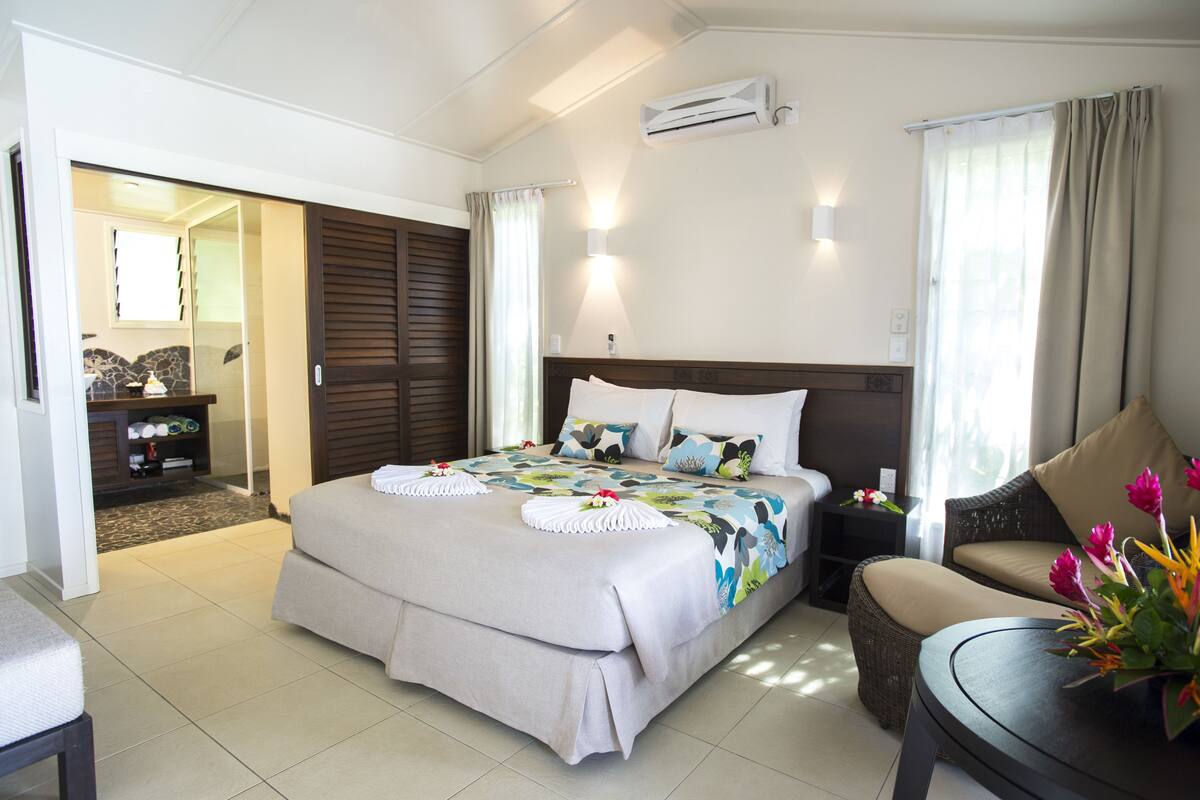 A hotel room with a large bed, a sofa, a small table, and a bathroom visible through an open door.