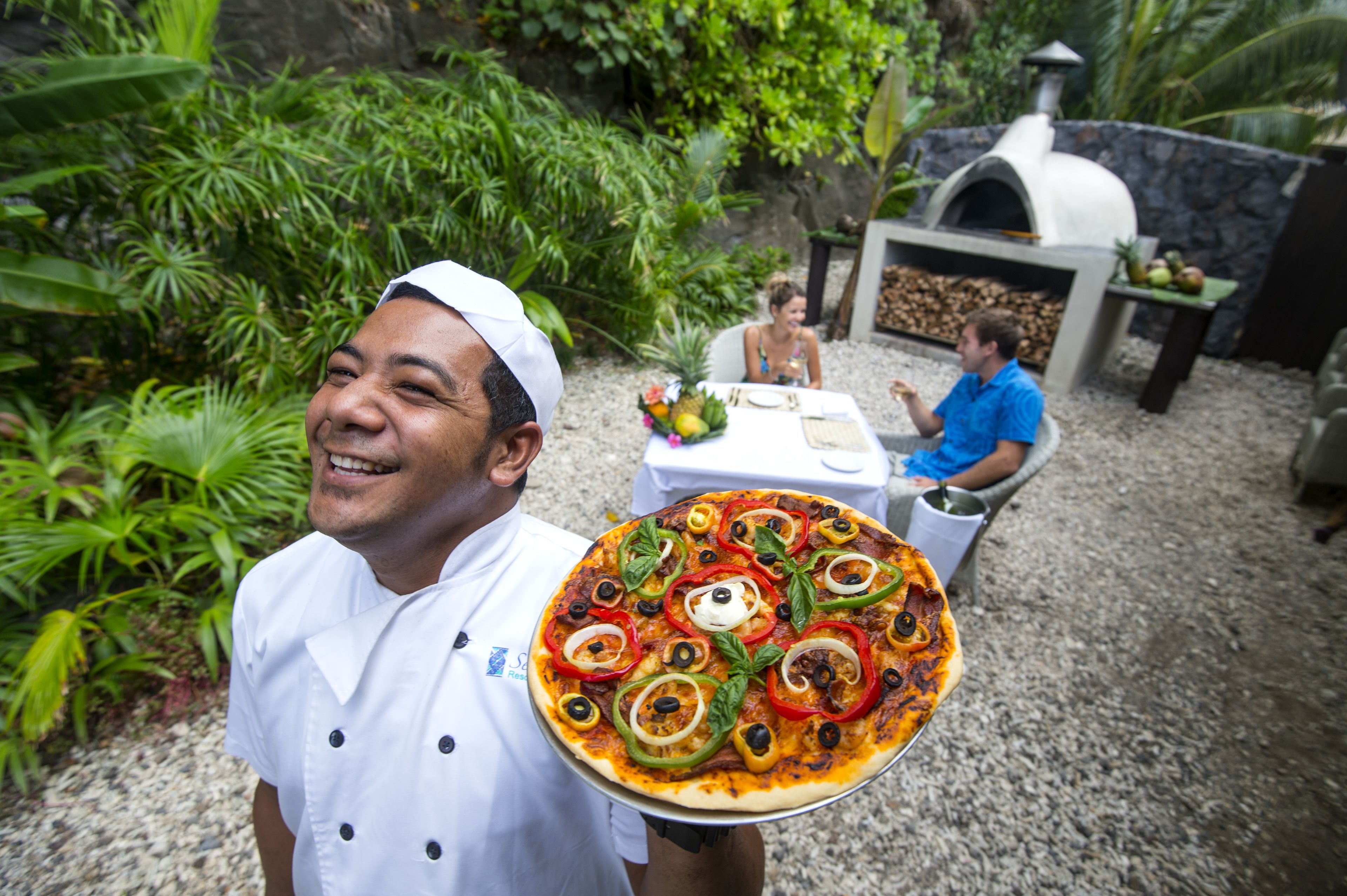 A chef holding a pizza with various toppings, a stone oven, and a dining area with a table and chairs.