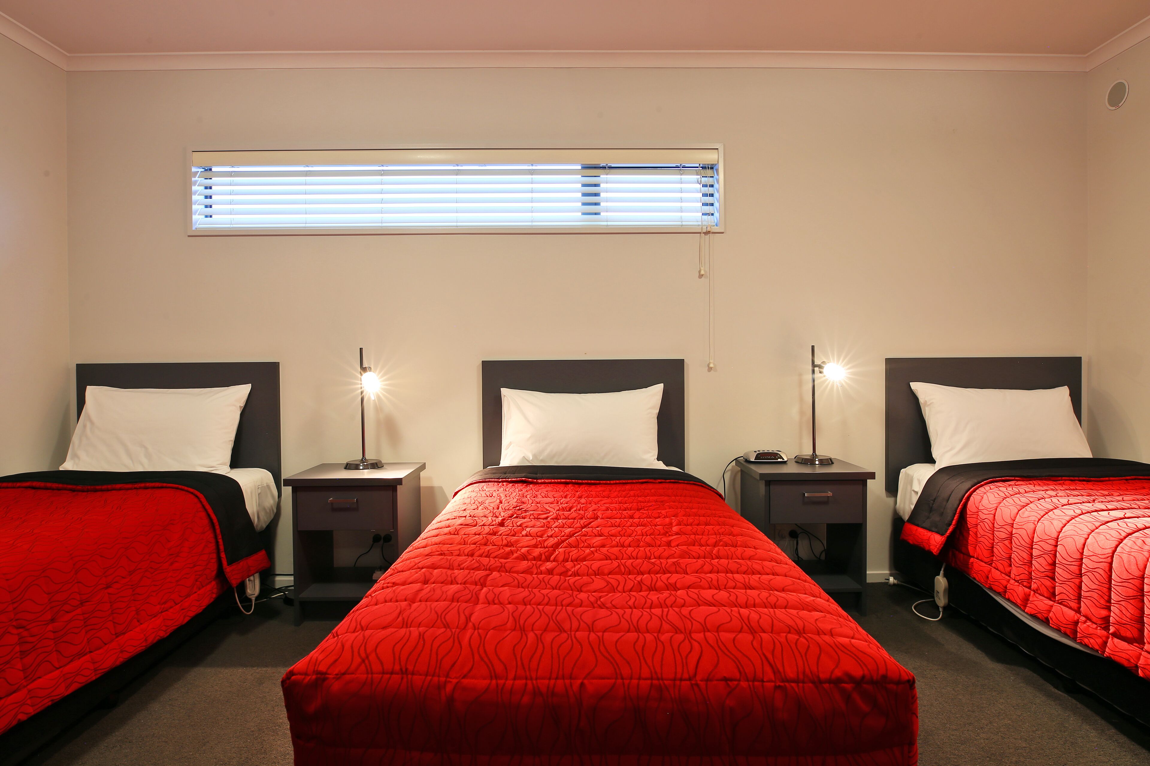 A hotel room with three single beds, each with a red bedspread and white pillows. There is a window with blinds, a bedside table with a lamp, and a small nightstand with a lamp.