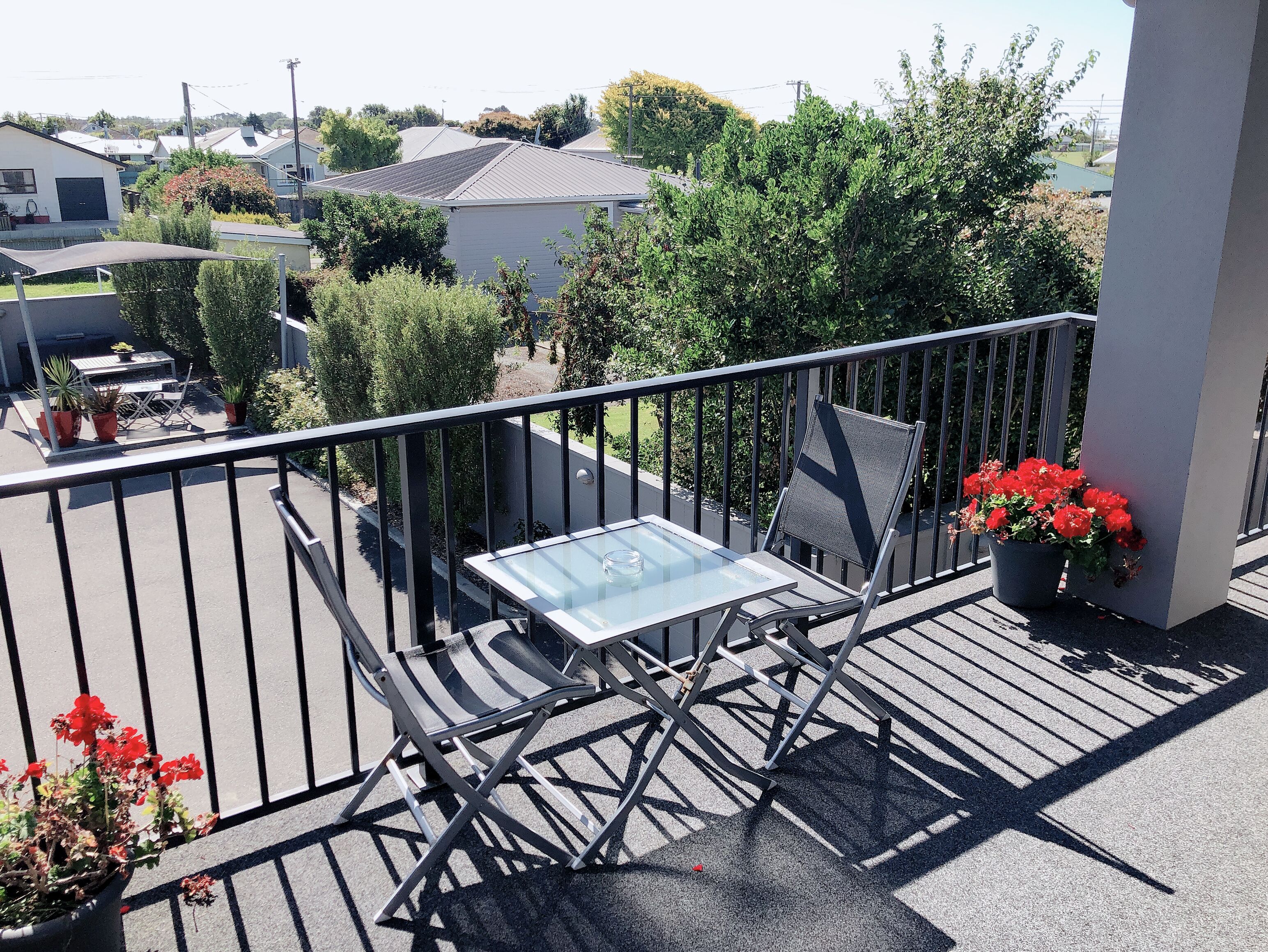 A balcony with a glass table and chairs, surrounded by potted plants and a view of a residential area.