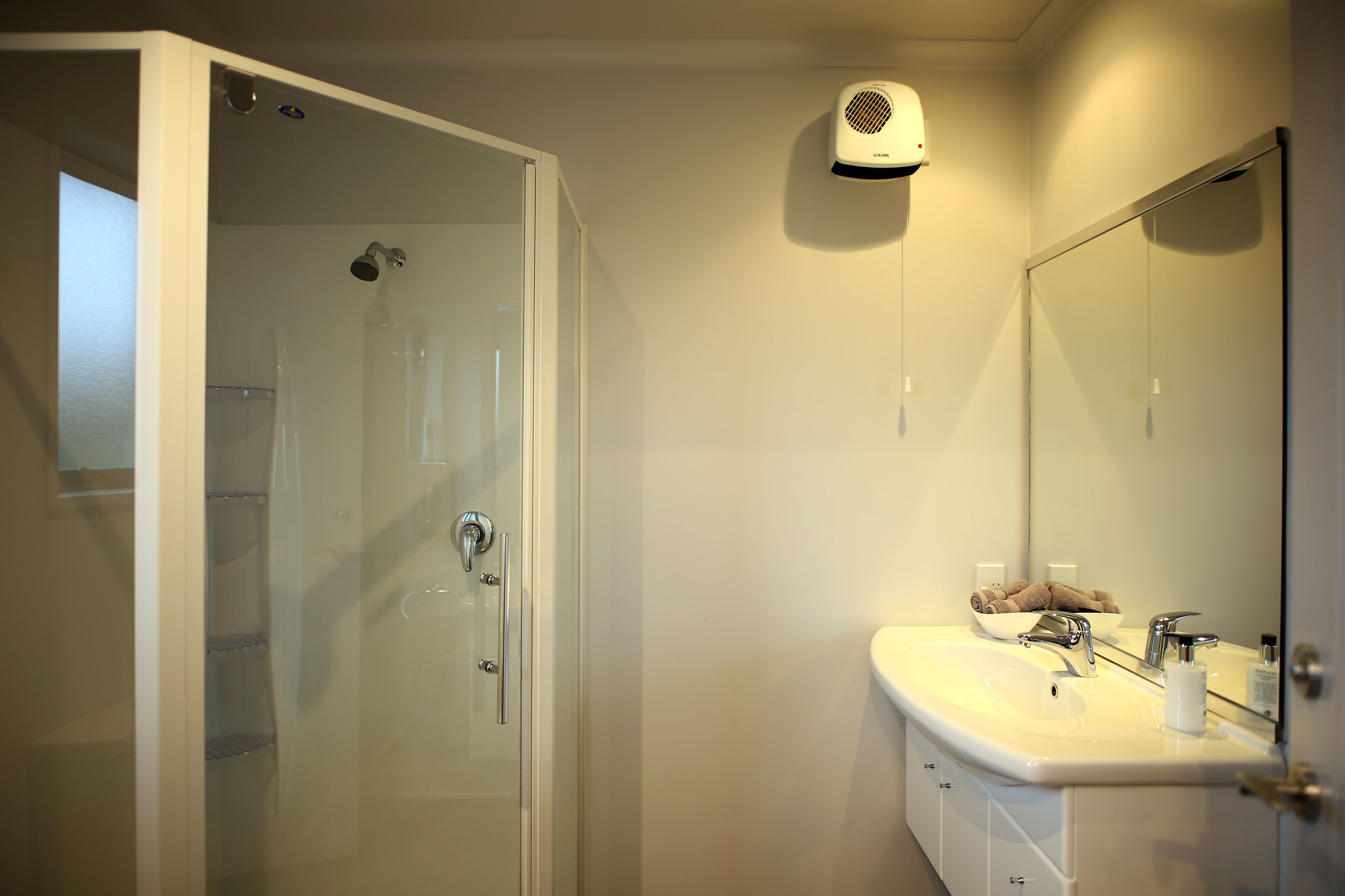 A bathroom with a glass shower enclosure, a white sink with two faucets, a mirror, and a hair dryer mounted on the wall.