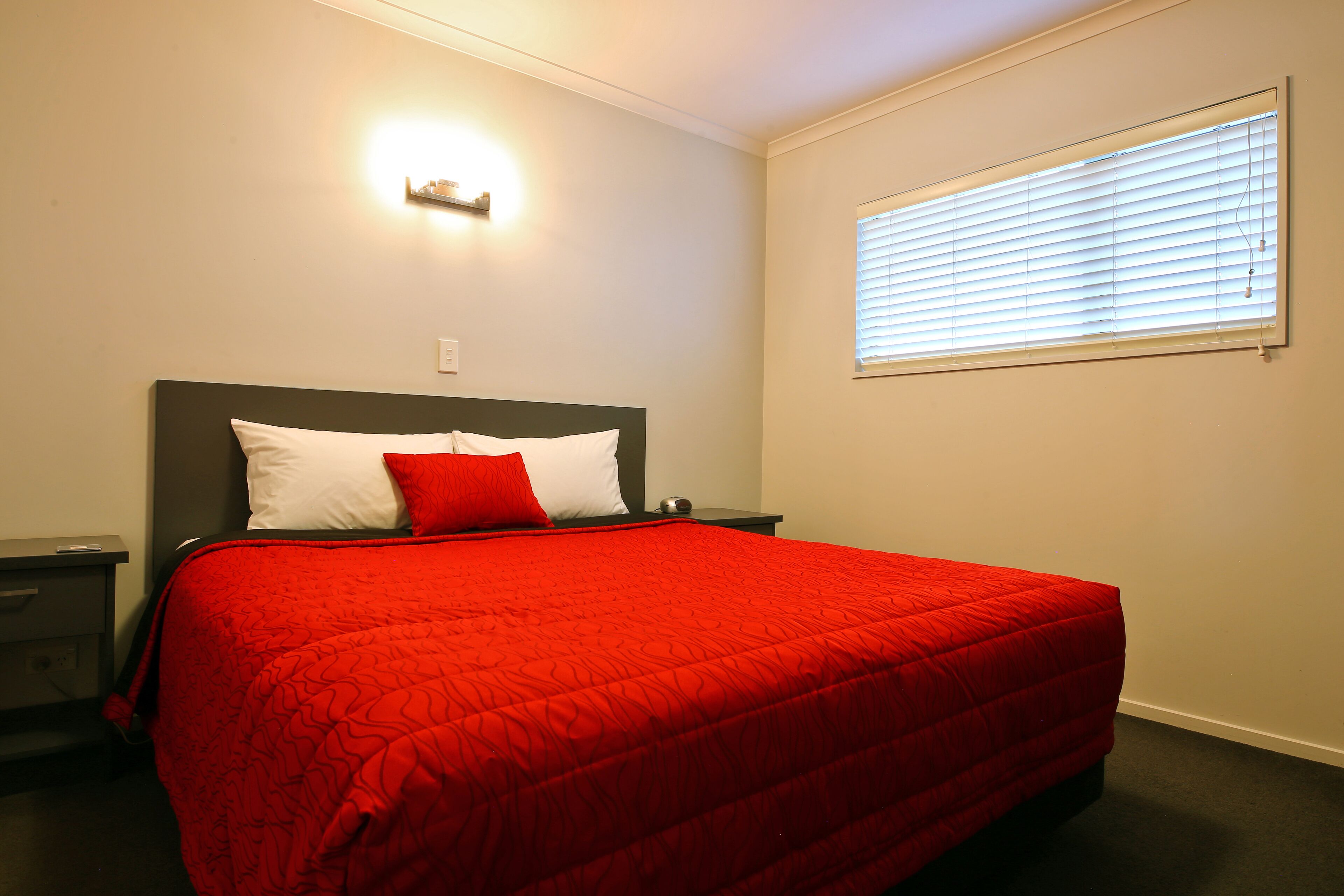 Un dormitorio con una cama, una mesita de noche, una lámpara de pared y una ventana con persianas.