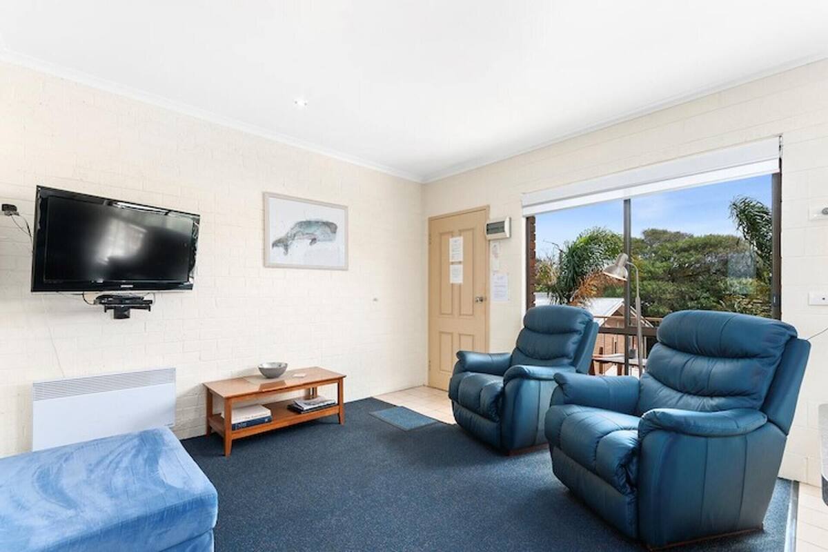 A room with a flat-screen TV mounted on the wall, a wooden coffee table, two blue leather armchairs, a blue sofa, and a large window offering a view of greenery outside.
