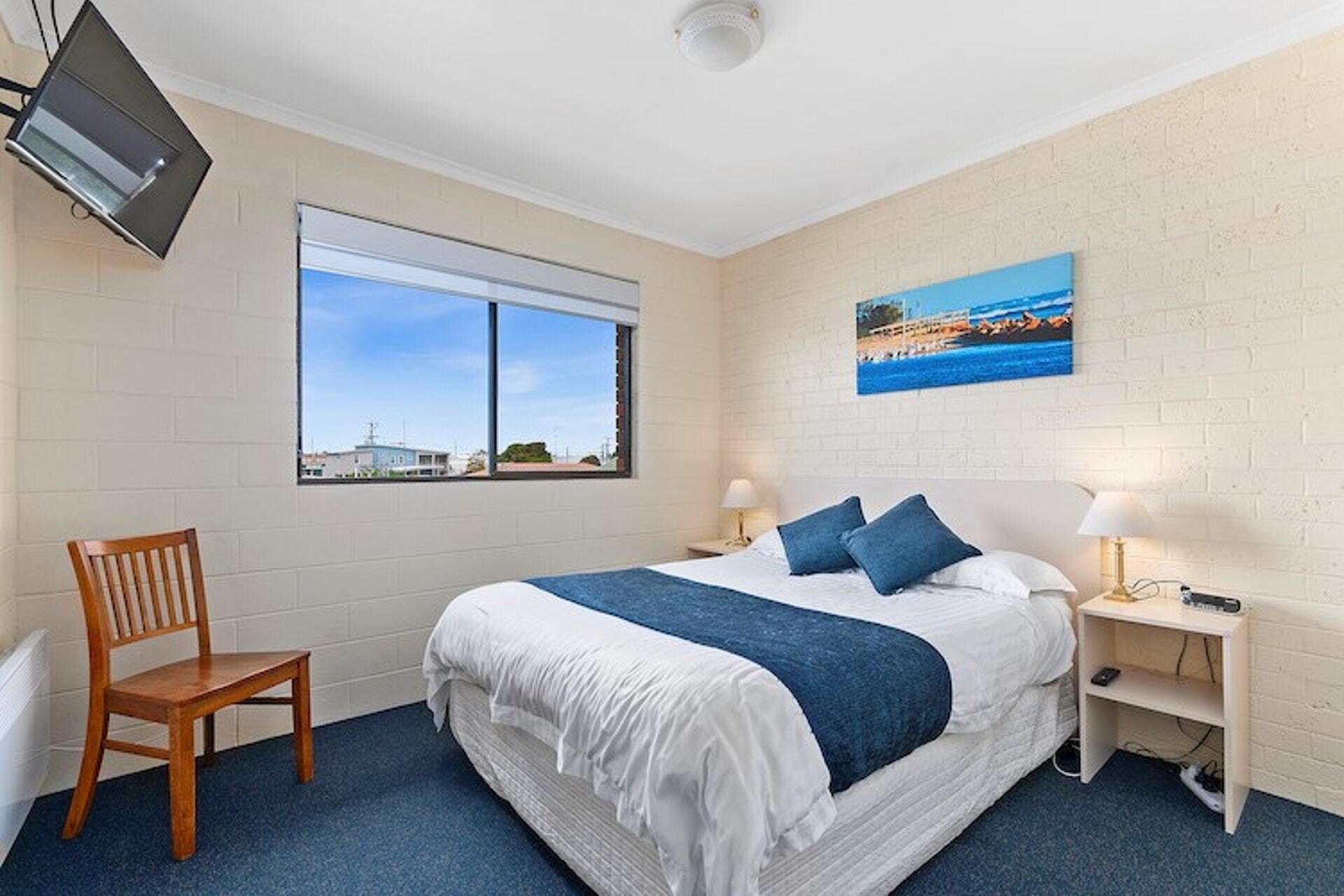 A hotel room with a large bed, two bedside tables, a chair, a TV mounted on the wall, and a window with a view of buildings.
