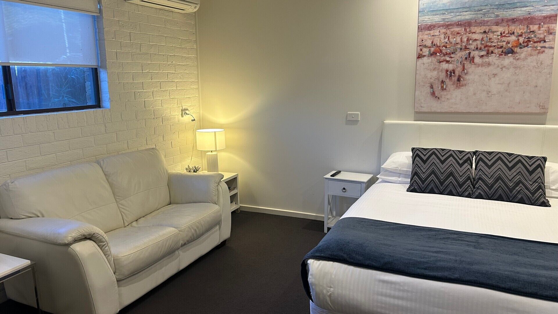 A hotel room with a white brick wall, a white sofa, a bed with a black and grey pillow, and a painting of a beach scene.