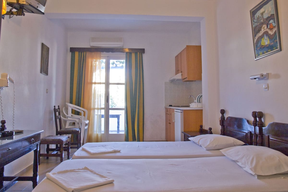 A hotel room with two beds, a desk, a chair, a small kitchenette, and a window with curtains.