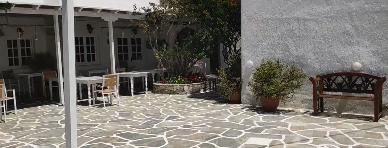 Een terras buiten met stenen vloer, witte tafels en stoelen, omringd door potplanten en een houten bank.