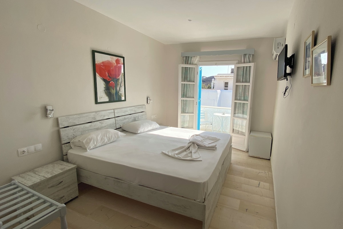 A bedroom with a bed, a nightstand, a radiator, a painting, and a view of the outside.
