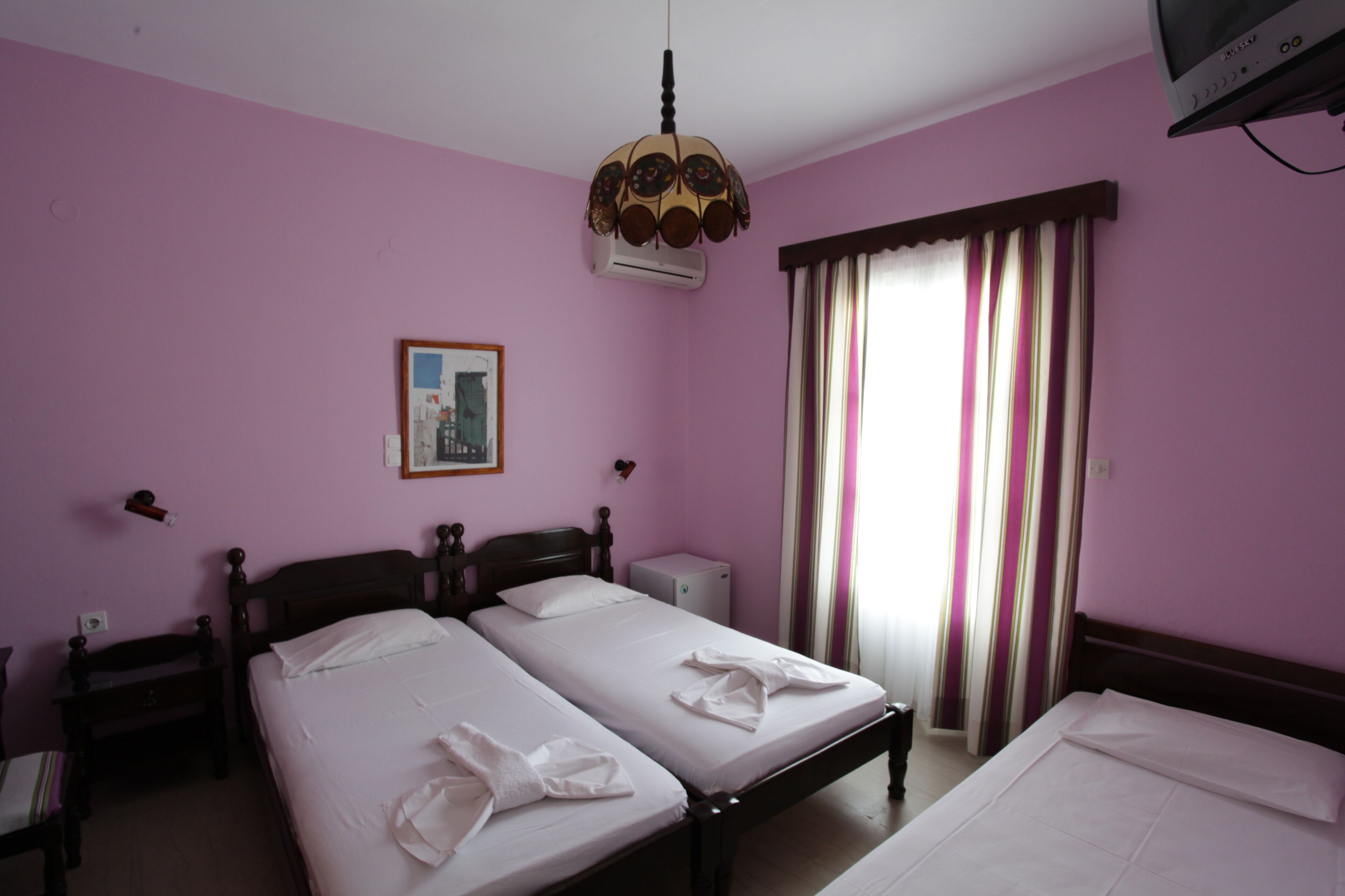 A room with two beds, a ceiling fan, a small table, a wall-mounted TV, and a window with curtains.