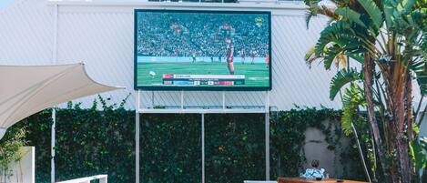 An outdoor seating area with a large screen displaying a sports event, surrounded by greenery and a wooden deck.