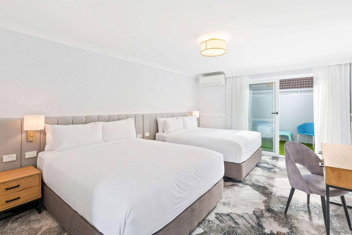 Une chambre d  ’hôtel avec deux lits, un bureau, une chaise, une table de chevet et une fenêtre avec des rideaux.