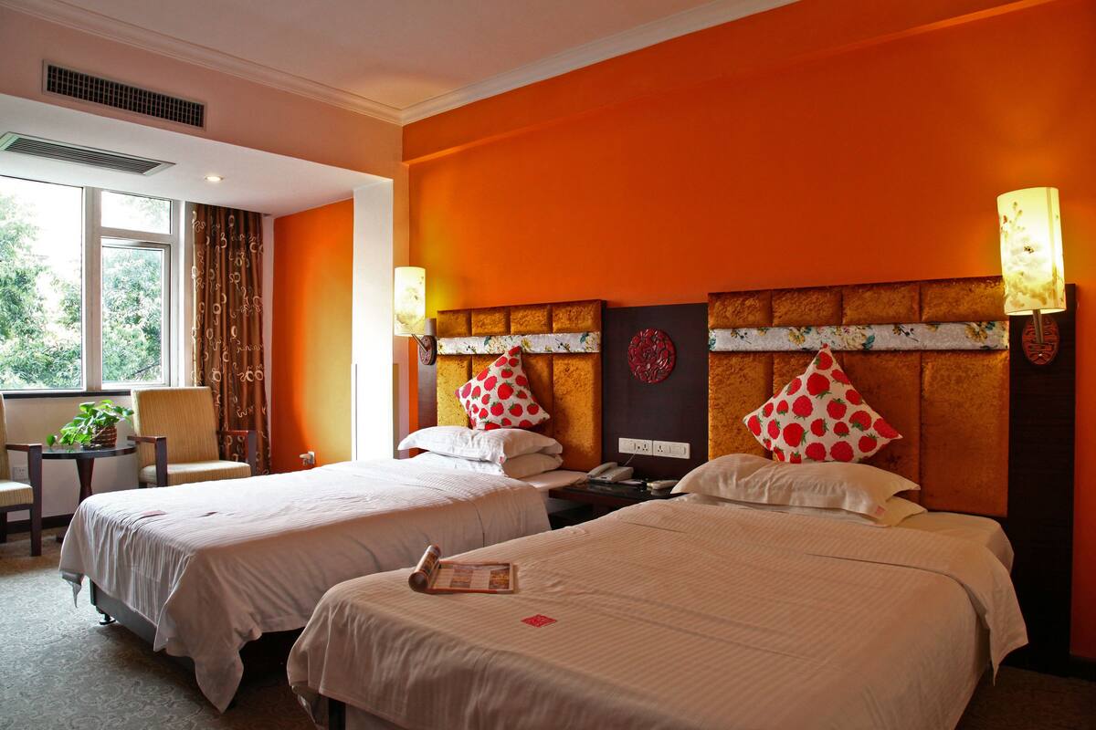 A hotel room with two beds, a desk, and a chair. There is a window with curtains, a plant, and a lamp on the wall.