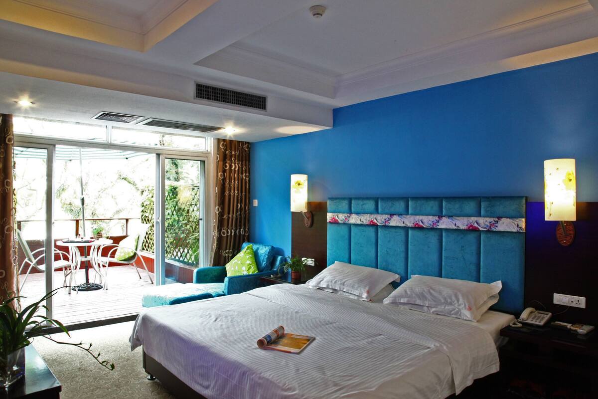 A hotel room with a large bed, a blue headboard, a balcony with a table and chairs, and a view of greenery outside.