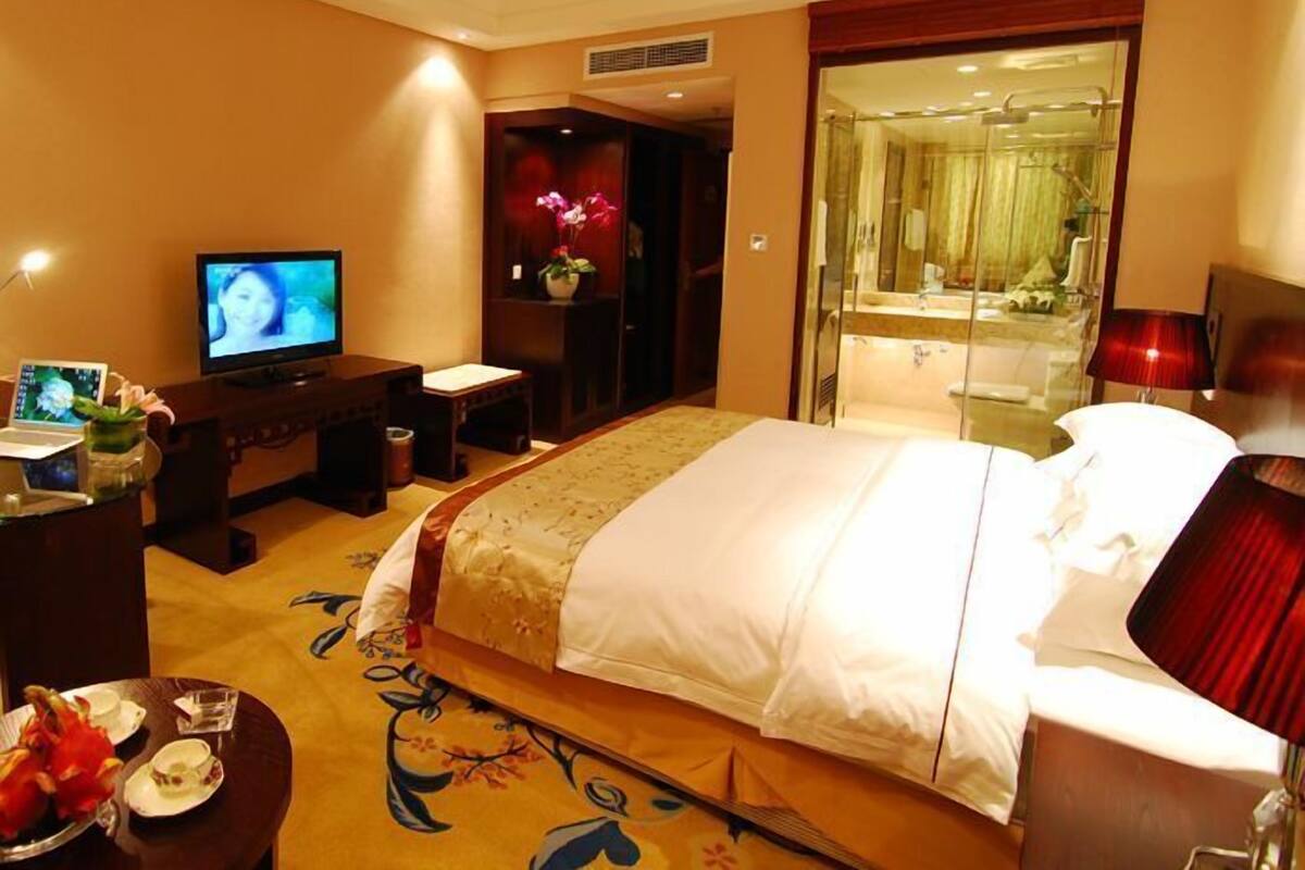A hotel room with a bed, a TV, a desk with a computer, a bathroom with a mirror, and a red lamp.