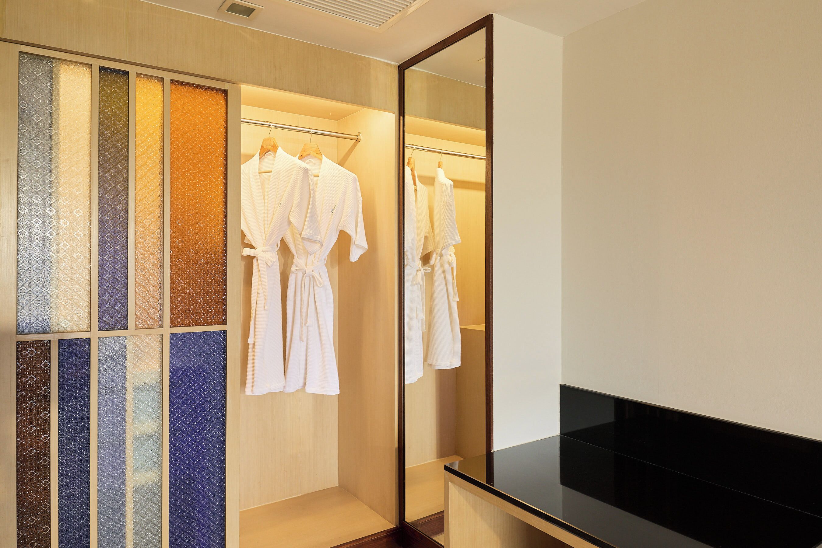Une chambre d’hôtel dotée d’un grand miroir, d’un comptoir noir et d’une armoire où sont suspendues des peignoirs.