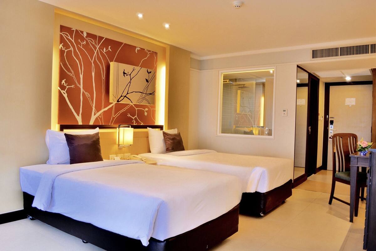 A hotel room with two beds, a large mirror, a wooden chair, and a wall-mounted artwork.