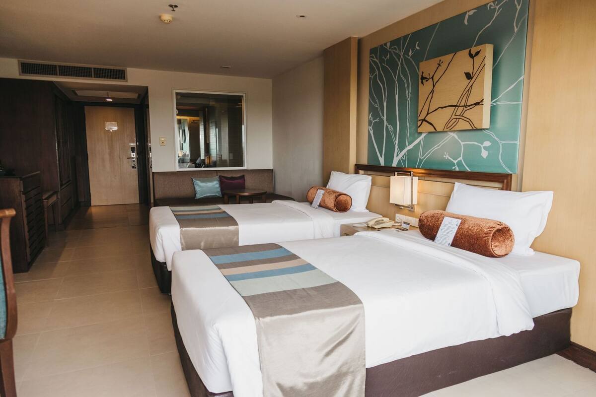A hotel room with two beds, a sitting area, a mirror, a wall art piece, and a view of the hallway.