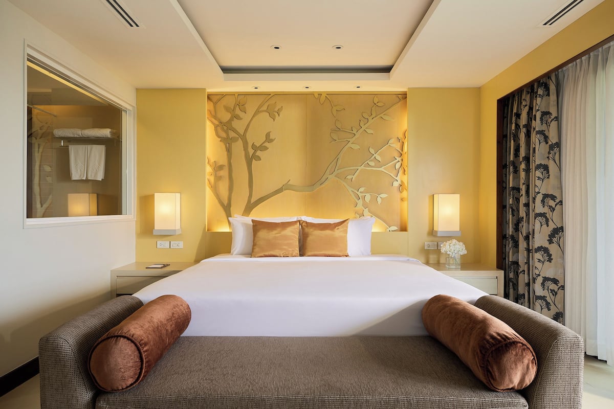 A hotel room with a large bed, two bedside lamps, a decorative wall panel, and a view of the outside through a window with curtains.