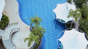 A large outdoor swimming pool with a curved design, surrounded by palm trees and shaded areas with umbrellas.