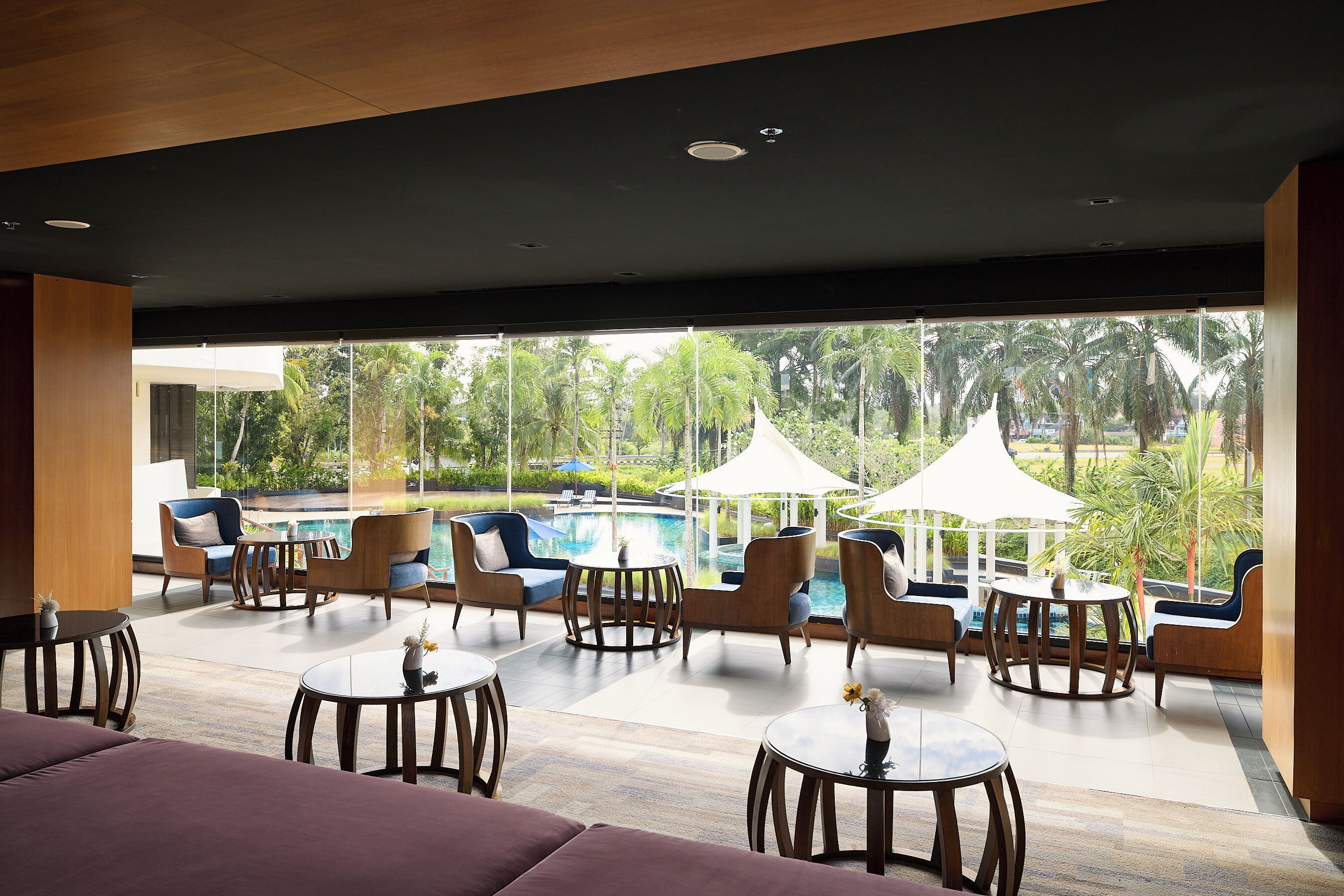 A modern lounge area with wooden furniture, a pool, and a view of palm trees.